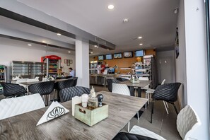 Een modern restaurant met verschillende zitmogelijkheden, waaronder tafels en stoelen. Er zijn meerdere schermen die content tonen aan de muur, en een Coca-Cola koelkast is zichtbaar.