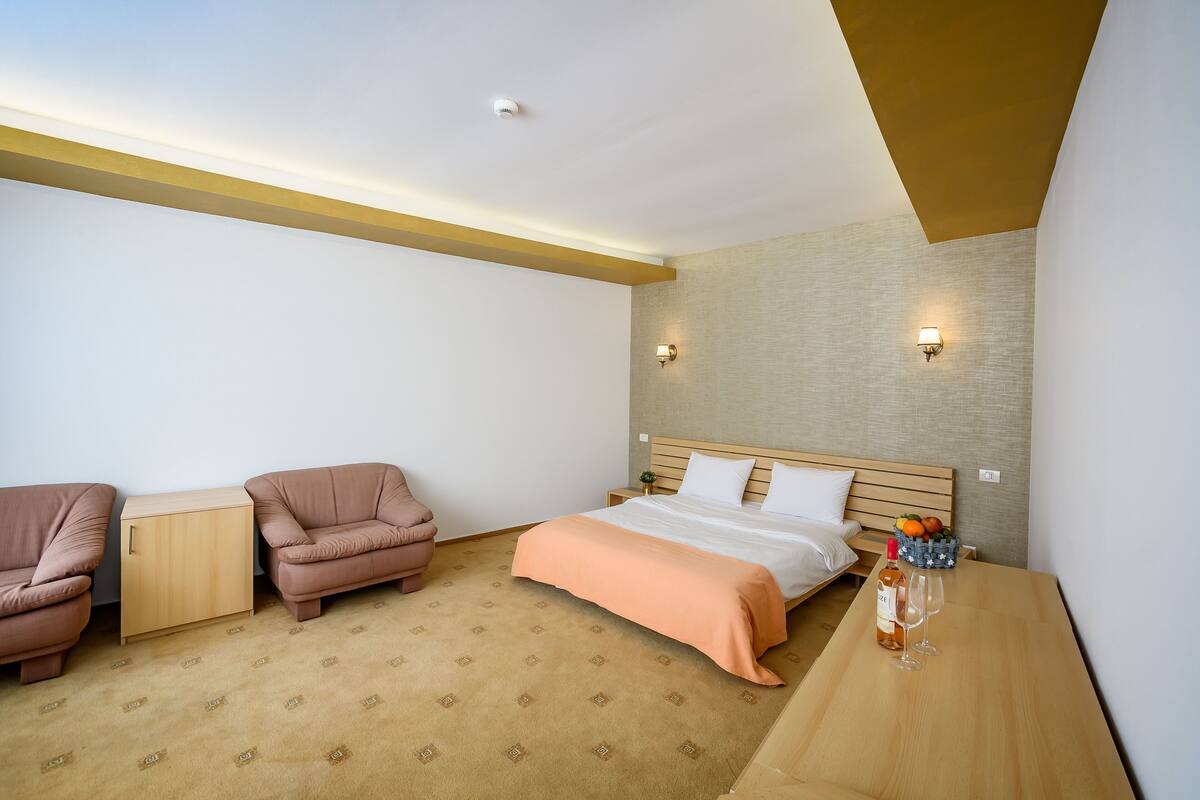 A hotel room with a bed, two armchairs, a wooden nightstand, and a table with a bottle and glasses.