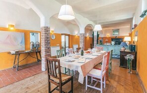 A dining area with a long table set for a meal, surrounded by chairs. There are hanging lights above the table, and the room has a rustic brick wall and wooden flooring.