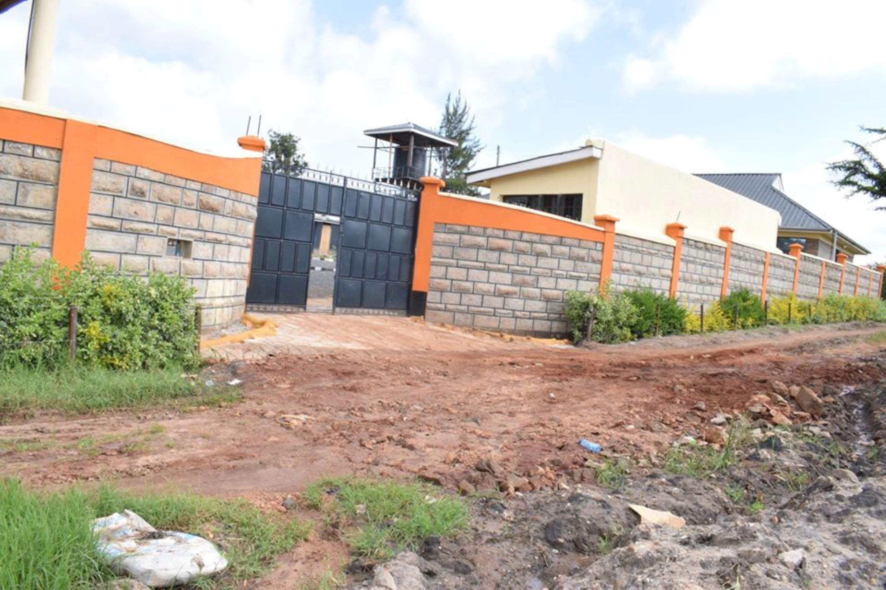 Uma entrada com portão e muro de tijolos, com detalhes em laranja, que leva a um edifício com telhado inclinado.