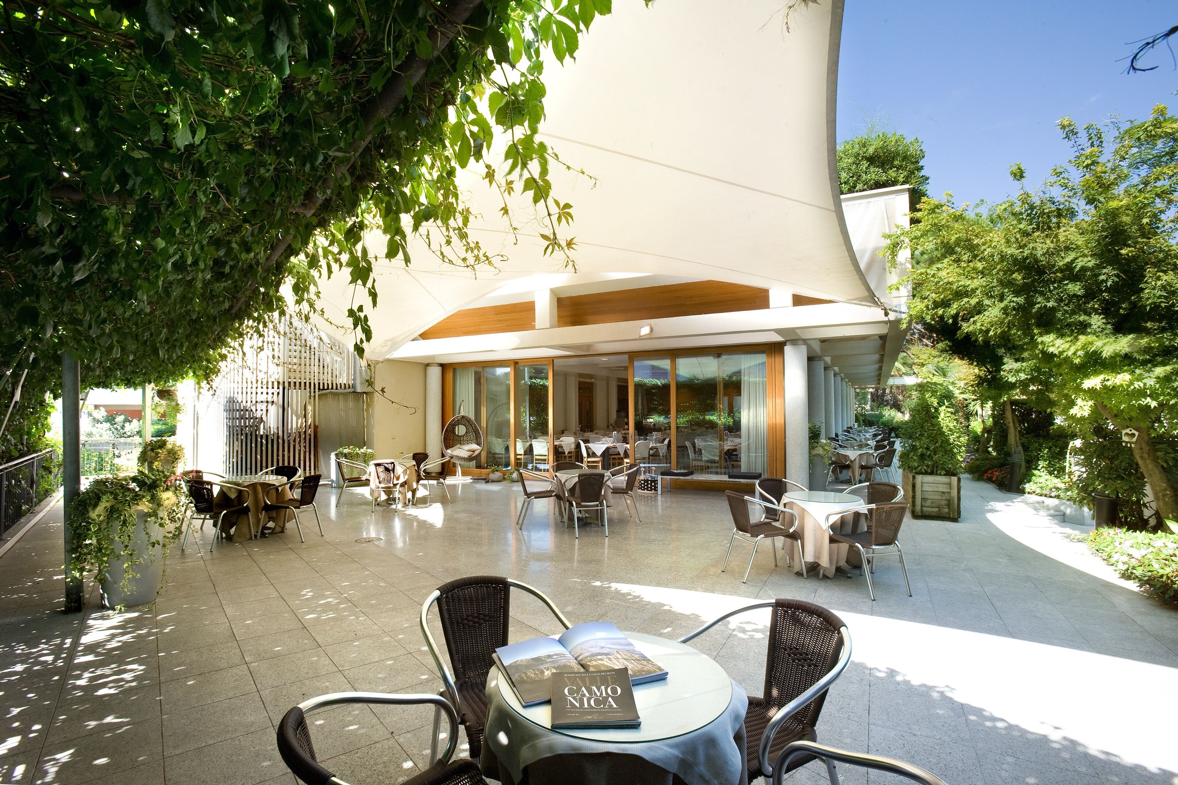 An outdoor cafe area with tables and chairs, surrounded by greenery and a modern building with large windows.