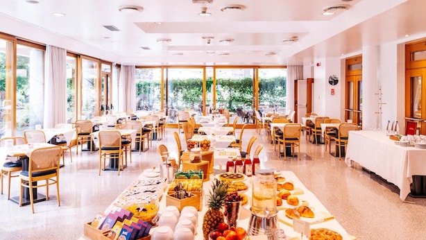 A spacious dining area with multiple tables and chairs, a buffet with various food items, and large windows offering a view of greenery outside. - Albergo Sorriso (Darfo Boario Terme)