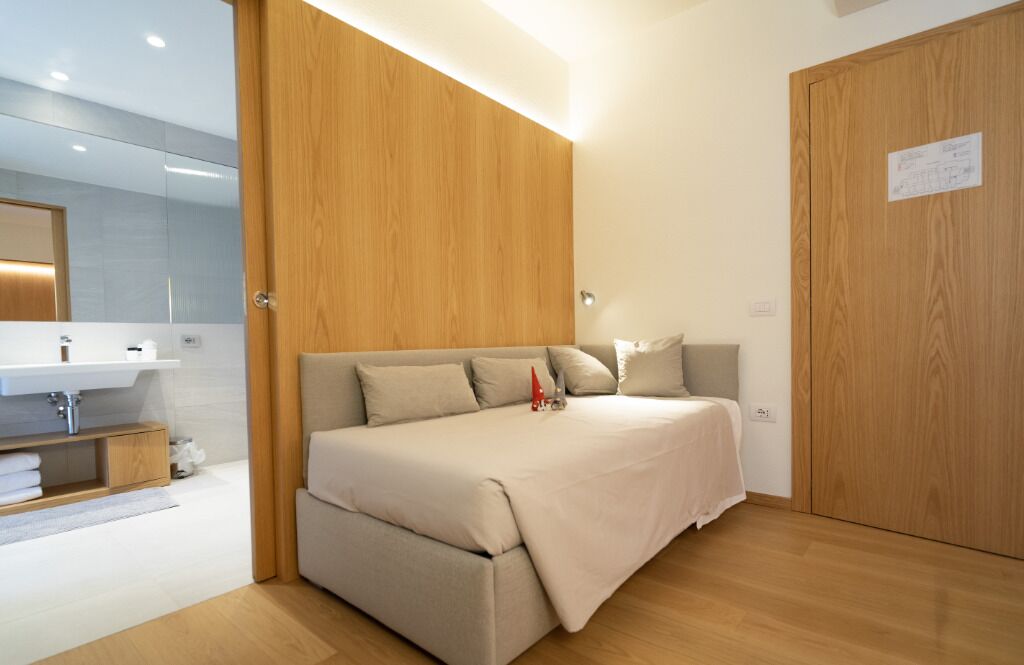 Ein modernes Schlafzimmer mit einem Holzschrank, einem Bett mit beigefarbener Bettwäsche und einem Badezimmer mit Waschbecken und Spiegel.