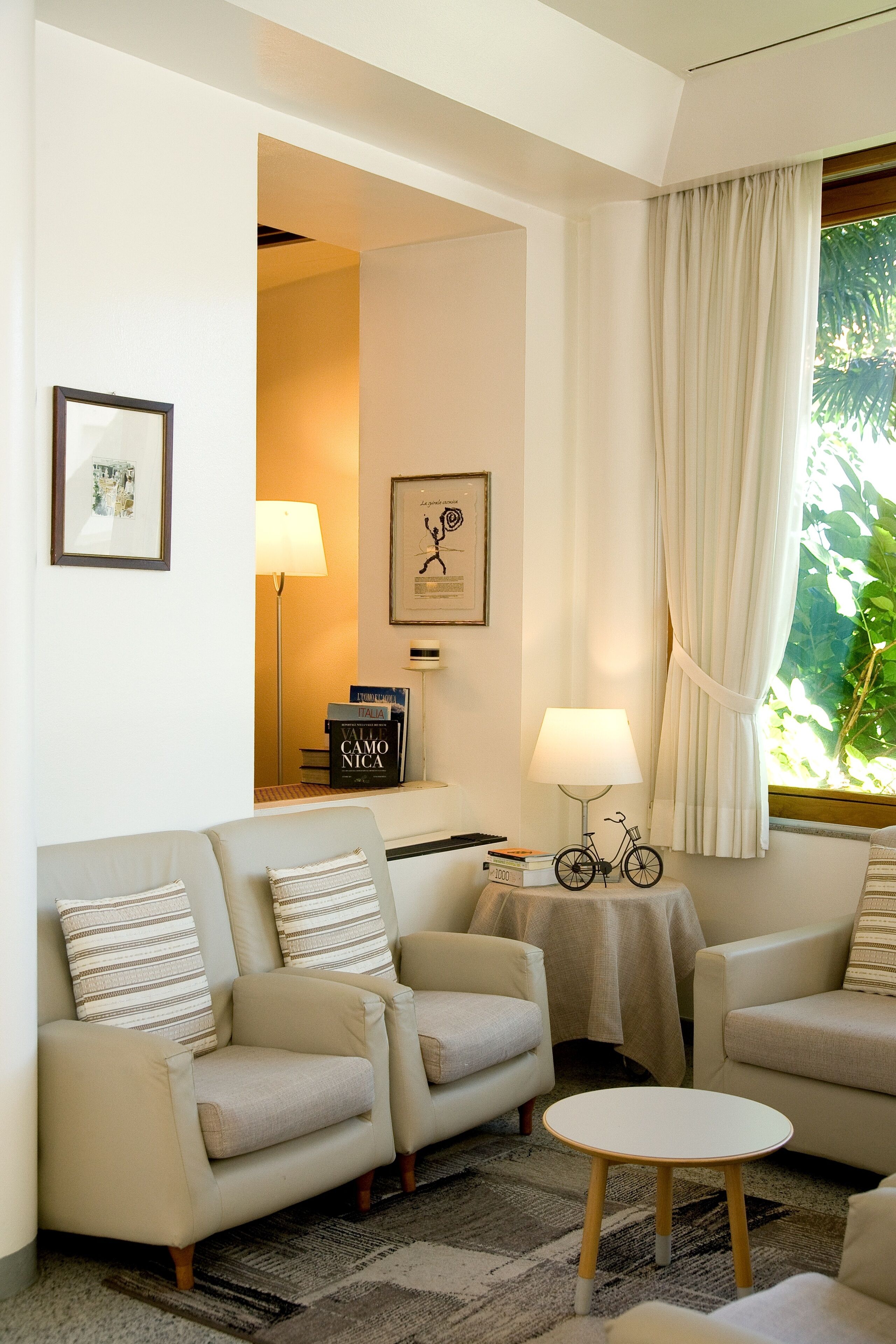 Un salon chaleureux avec un canapé, des fauteuils, une petite table ronde et une lampe. Des tableaux encadrés ornent le mur, et une fenêtre est agrémentée de rideaux.