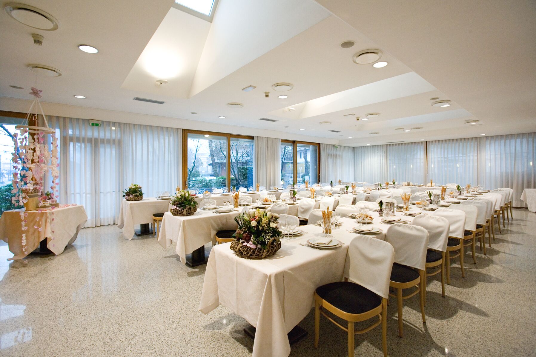 Une salle de banquet avec de nombreuses tables rondes dressées pour le repas, ornées de nappes blanches et de compositions florales.