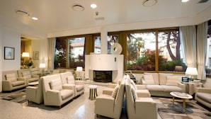 A spacious living room with a fireplace, multiple seating areas, and large windows offering views of greenery and buildings. - Albergo Sorriso (Darfo Boario Terme)