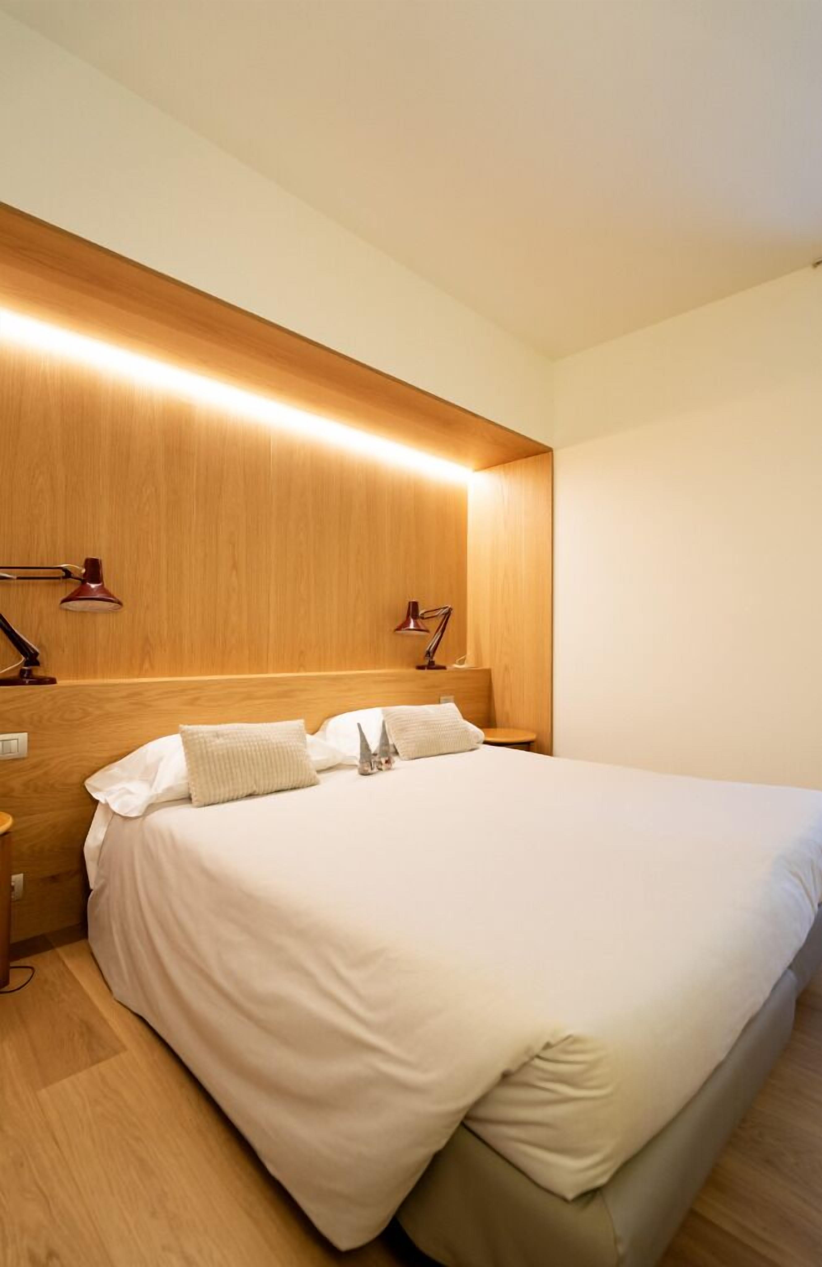 A modern hotel room with a wooden headboard, a large bed with white linens, two bedside lamps, and a nightstand.