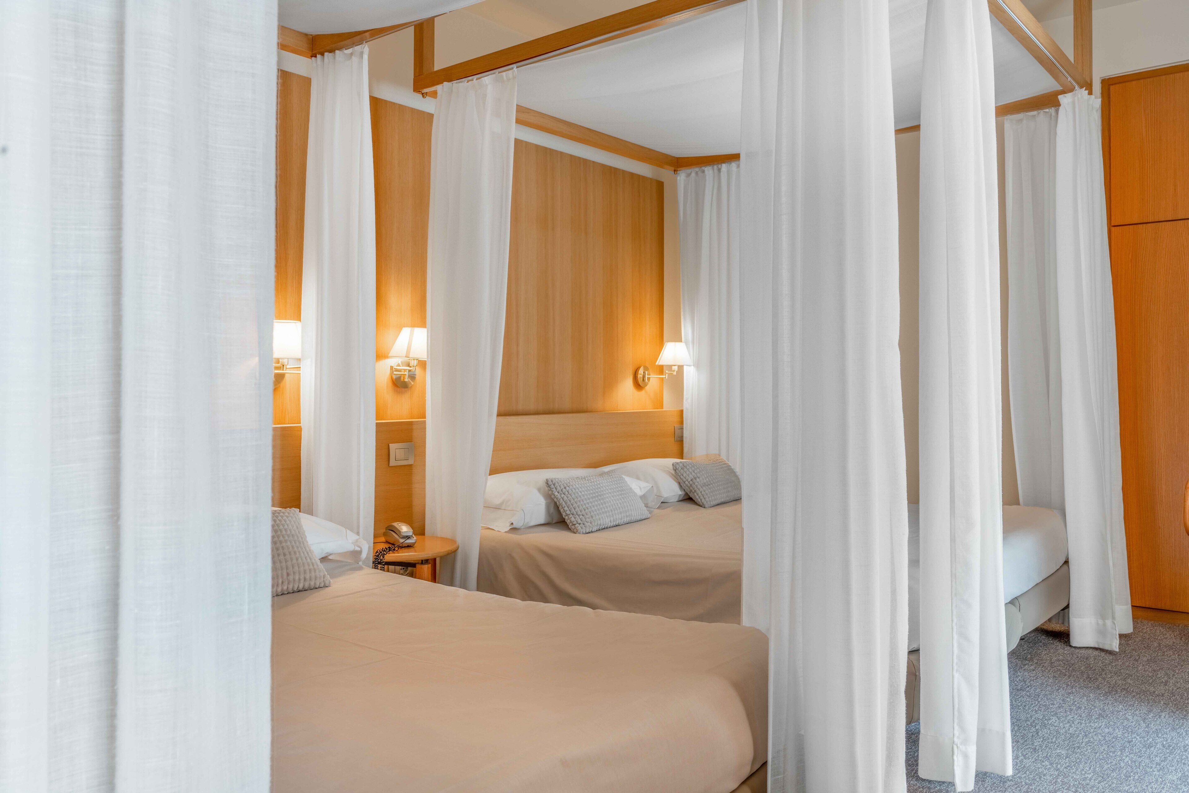 A hotel room with two beds, wooden headboards, white curtains, bedside lamps, and a wooden wardrobe.