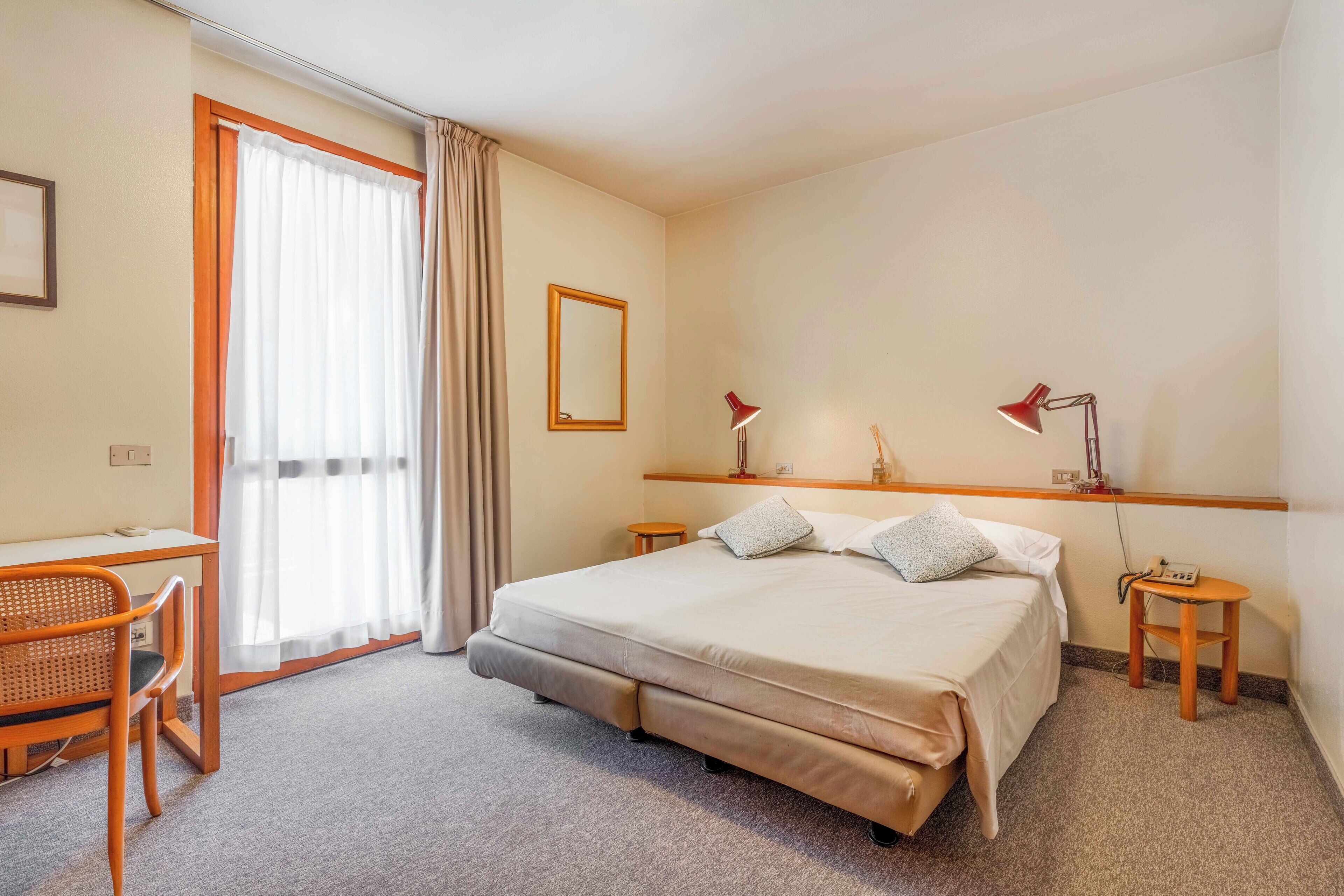 Een hotelkamer met een bed, een bureau, een stoel, twee nachtkastjes met lampen, een spiegel en een raam met gordijnen.