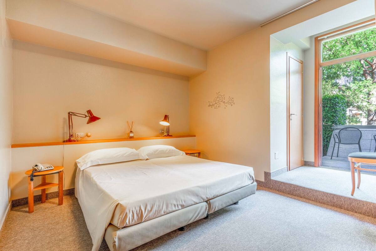 Een hotelkamer met een bed, nachtlampjes, een klein tafeltje met een telefoon en uitzicht op het omliggende landschap door een schuifdeur.
