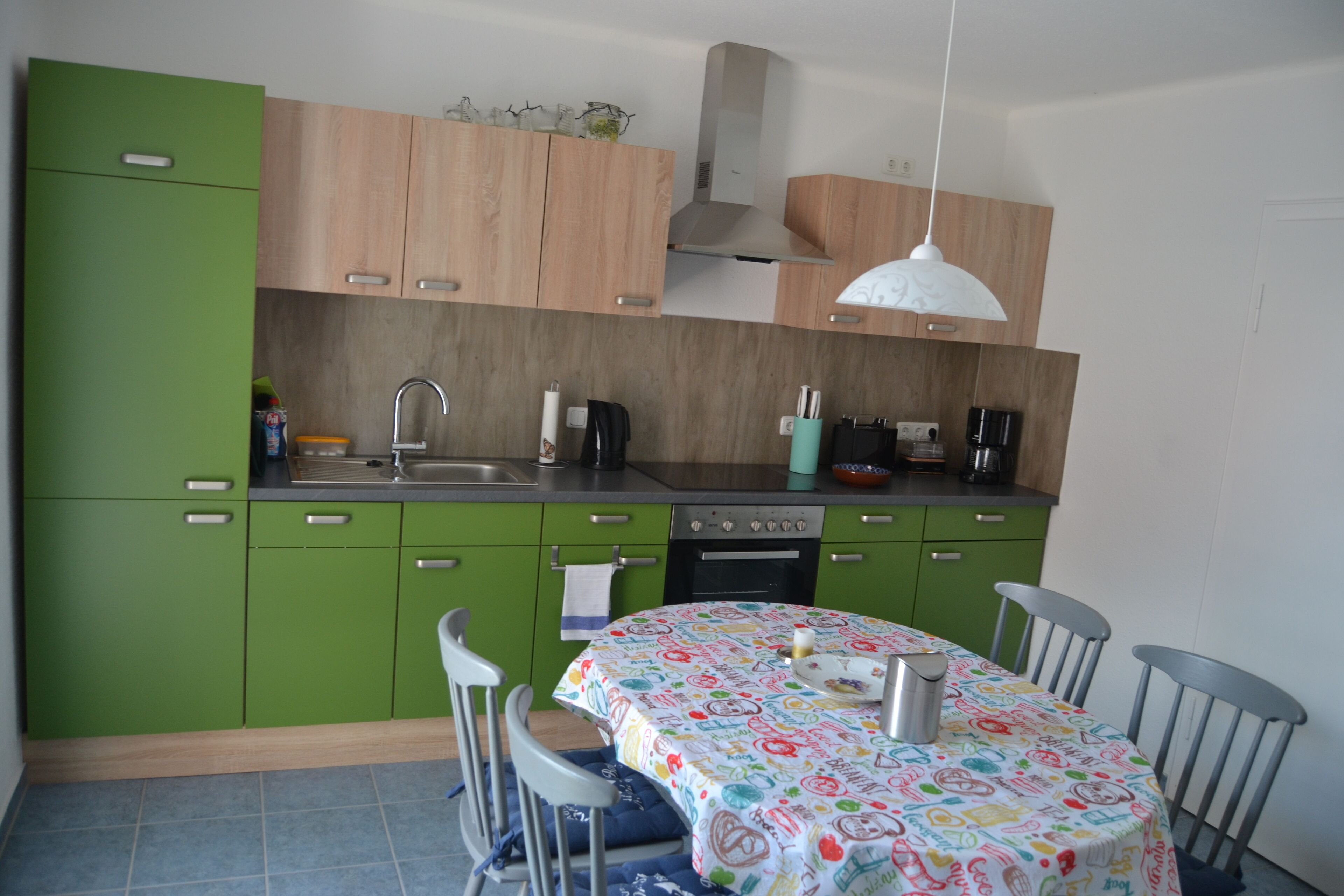 Une cuisine avec des armoires vertes, un plan de travail en bois, une cuisinière et une table recouverte d’une nappe colorée.