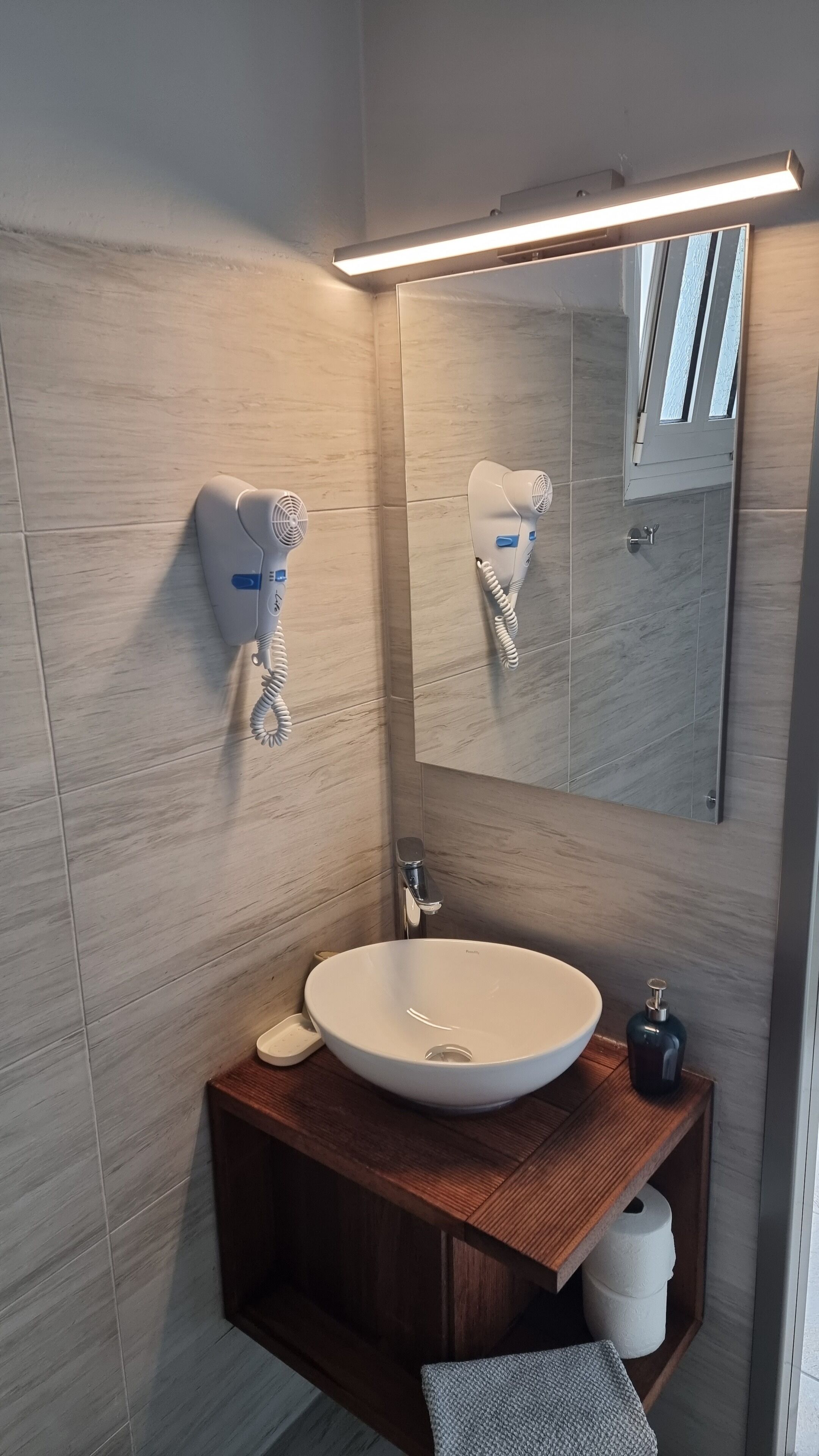Een moderne badkamer met een grote spiegel, een witte wastafel, een zeepdispenser en een houten opbergmeubel.
