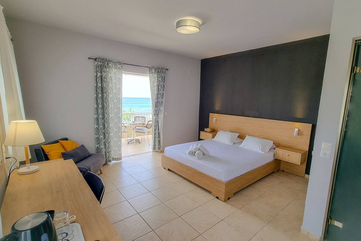 A hotel room with a bed, bedside tables, a desk with a lamp, a chair, and a view of the sea through a window.