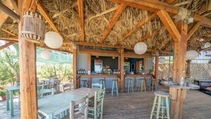 A wooden deck with tables and chairs, a bar area, and a thatched roof. - Chloe Apartments (Kefalonia)
