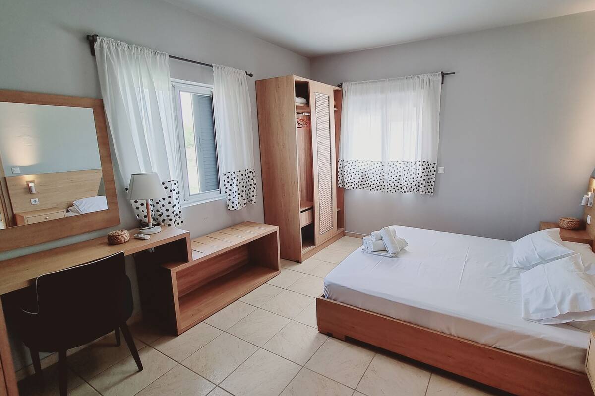 A hotel room with a wooden bed, a desk with a lamp, a wardrobe, and a window with curtains.