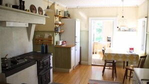 A compact kitchen with a stove, refrigerator, and wooden shelves. A dining area with a table and chairs is visible through an open door. - Holiday home Törnamåla Kilen (Tingsryd)