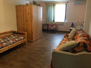 A room with a wooden wardrobe, a sofa with patterned cushions, a bed with a quilt, a chair, and a window with curtains.