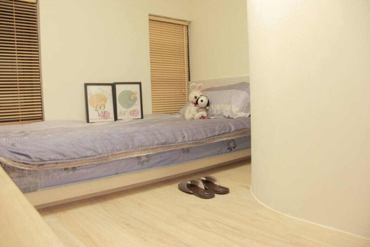 A single bed with a mattress, pillows, and a teddy bear. There are two framed pictures on the wall, a pair of slippers on the floor, and a window with blinds.