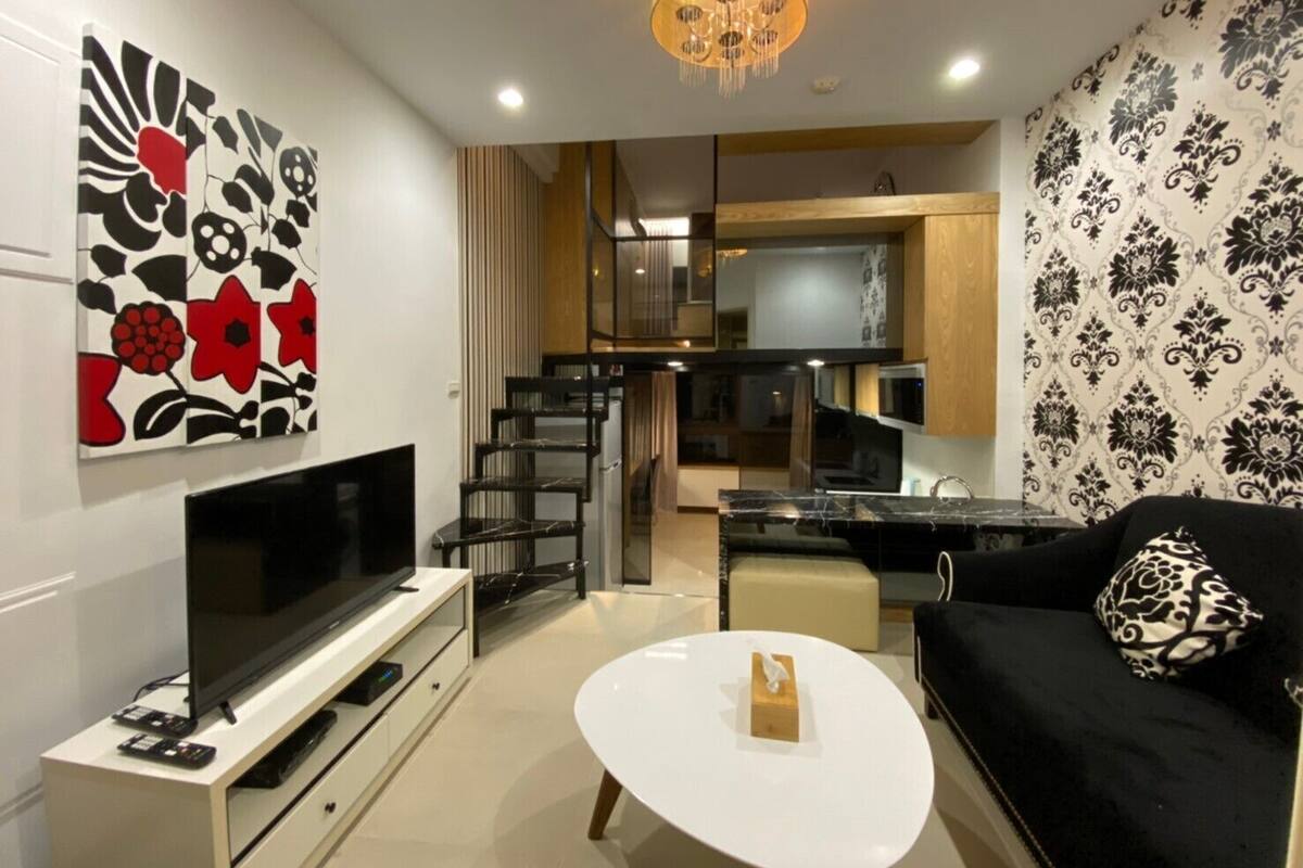 A modern living room with a flat-screen TV, a white coffee table, and a black sofa with a patterned pillow. A staircase leads to a kitchen area with a microwave and a wooden cabinet.