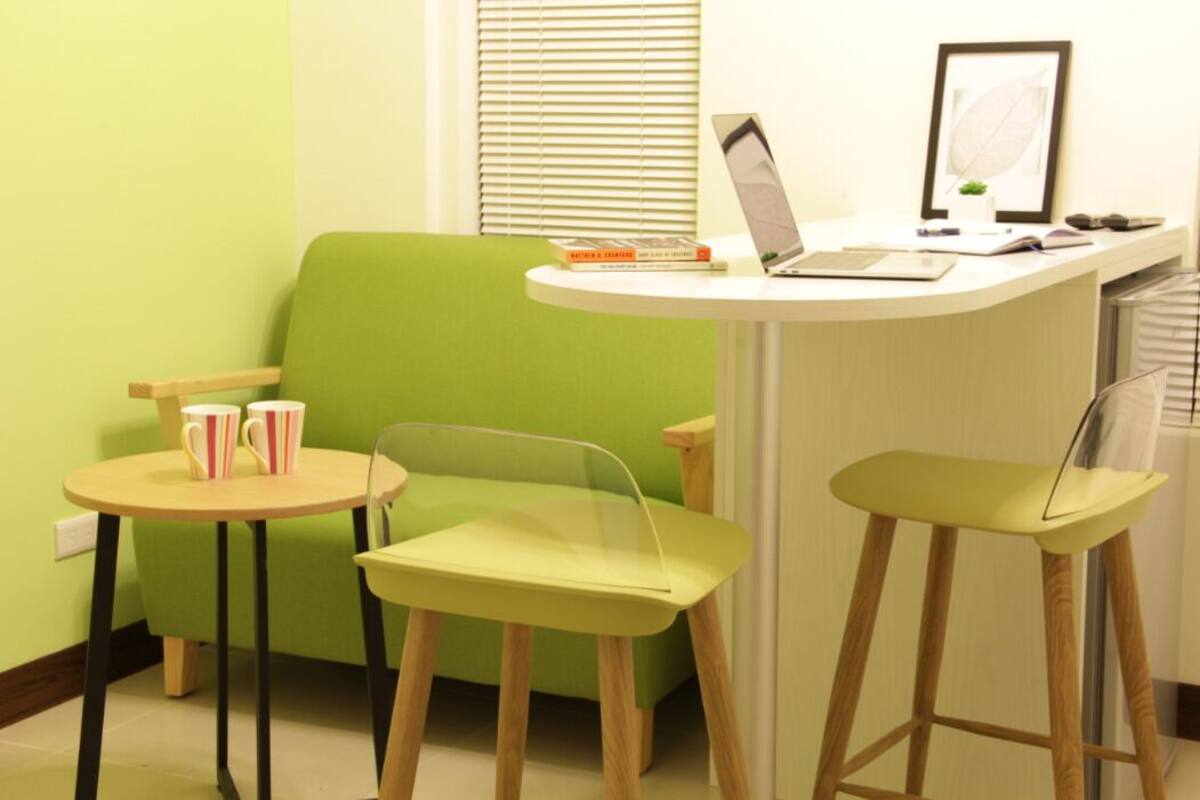 A small, brightly colored room with a green sofa, two chairs, a small table, and a laptop on a white desk.