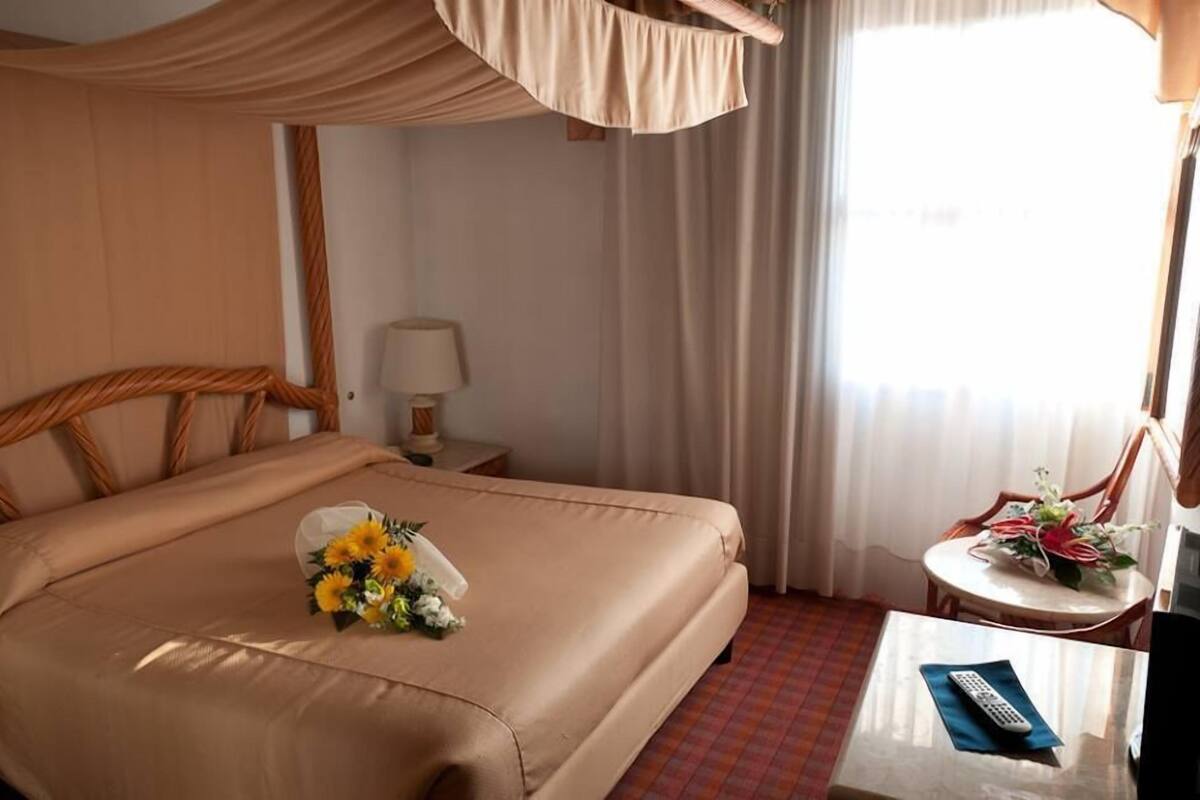 A hotel room with a bed, a small table with a remote control, a chair with a floral arrangement, and a window with curtains.