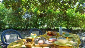An outdoor dining table with a floral tablecloth, surrounded by chairs, set with bread, bowls, and a bottle.