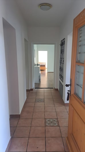 Ein Flur mit Fliesenboden, einer Holztür und Blick in einen Raum mit Bett und Fenster.