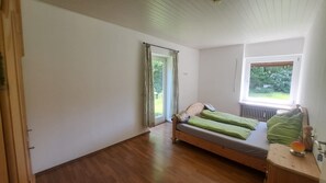 Quarto com piso de madeira, cama com roupa de cama verde, janela com vista para áreas verdes e radiador.
