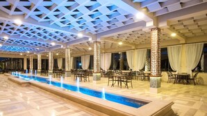 A spacious indoor pool area with a long, narrow pool, surrounded by tables and chairs, and a ceiling with a geometric pattern. - One bedroom "Beach Haven" in 5-stars resort with pool and private beach (Sahl Hasheesh)