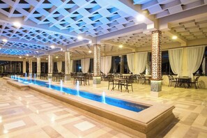 Amplia zona de piscina interior con una piscina larga y estrecha, rodeada de mesas y sillas, y un techo con diseño geométrico.