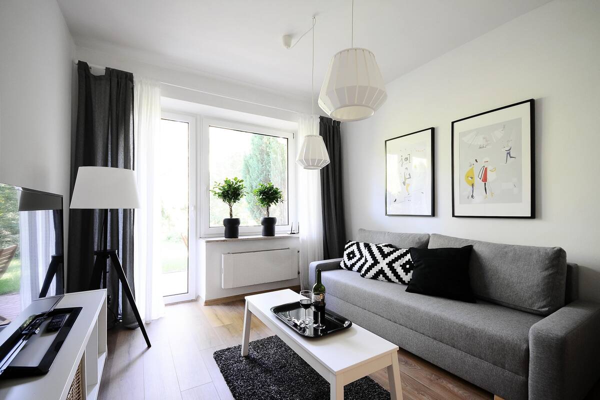 Een moderne woonkamer met een grijze bank, een witte salontafel, een vloerlamp en een groot raam met gordijnen.