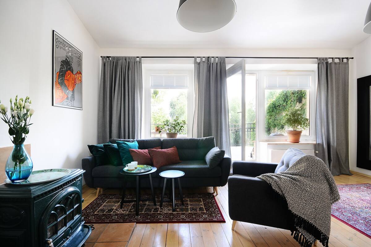 Een woonkamer met een bank, salontafel en een potplant. Er is een raam met gordijnen, een open haard en een televisie.