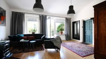 A living room with a sofa, a small table, a chair, and a rug. There are two windows with curtains, a door, and a potted plant.