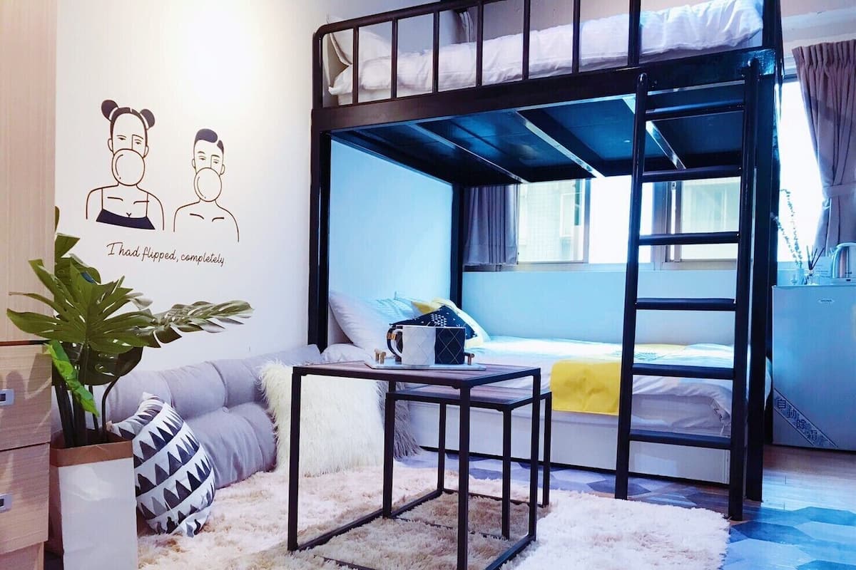 A dorm-style room with bunk beds, a small table, a plant, and a window.