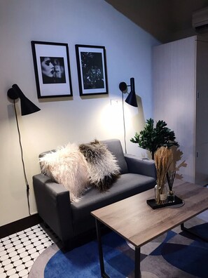Una sala de estar moderna con un sofá gris, una mesa de centro de madera y dos lámparas de pie negras. Hay dos cuadros enmarcados en la pared y una planta en maceta sobre la mesa.