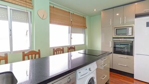 Een kleine keuken met een vaatwasser, wasmachine, magnetron en oven. Er staan houten stoelen en een tafel, en er hangt een klok aan de muur.