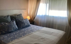 A neatly made bed with patterned bedding, a bedside lamp, and sheer curtains. - Rubens Center apartment (Sevilla)