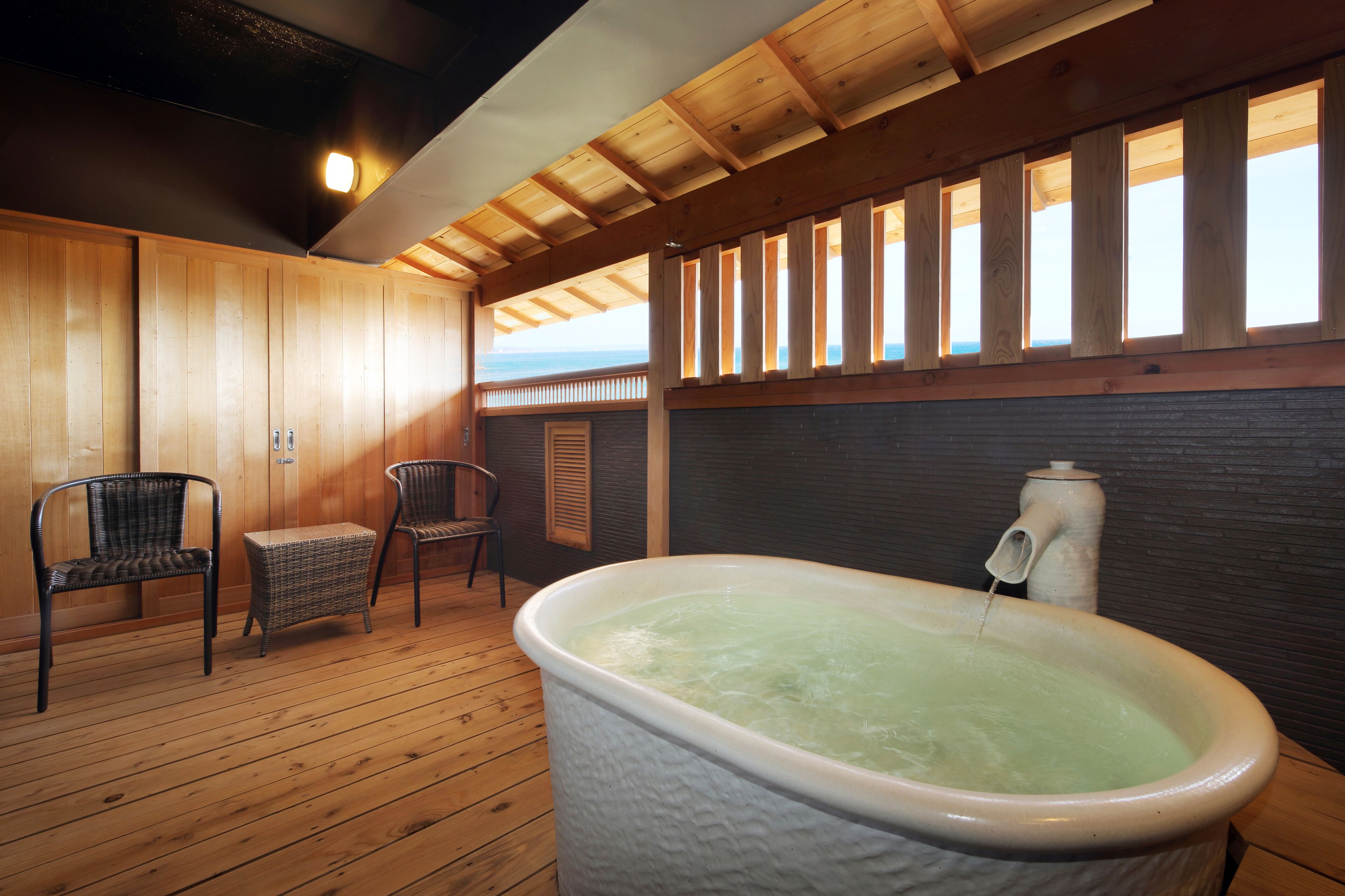 A wooden-floored room with a large bathtub, two wicker chairs, and a view of the ocean through a window.