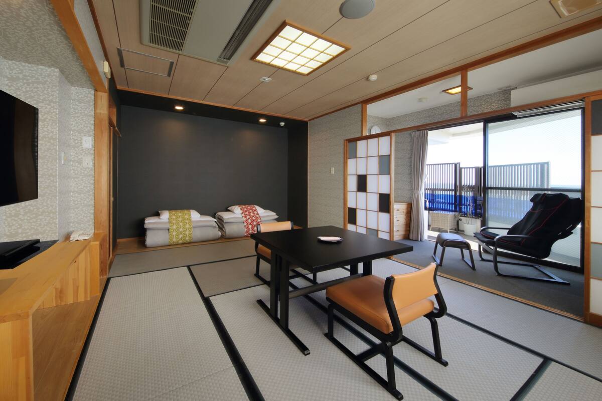 A modern hotel room with tatami flooring, a black table with chairs, a sofa with pillows, and a sliding glass door leading to a balcony.