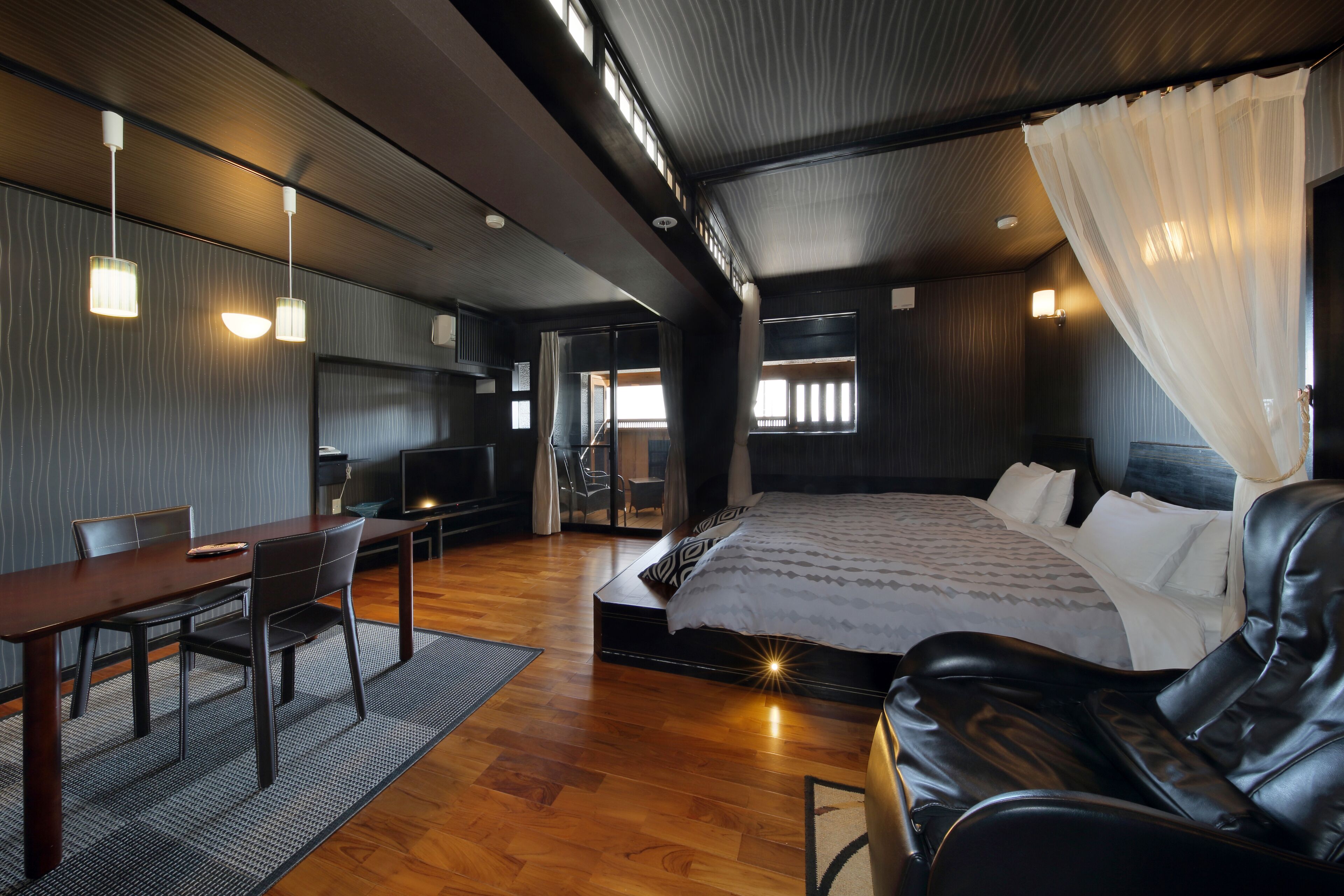 Una habitación de hotel moderna con piso de madera, una cama grande con ropa blanca, un sofá de cuero, una mesa de comedor con sillas e iluminación colgante.