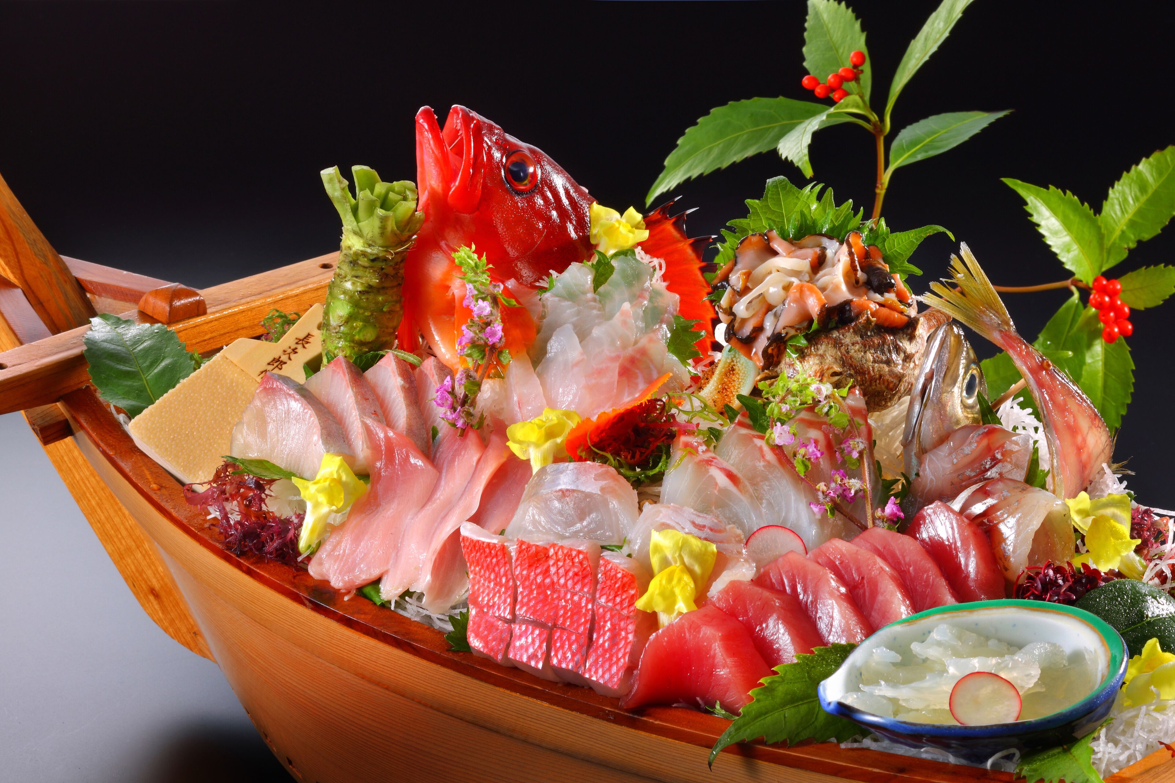 Un barco de madera lleno de diferentes tipos de sashimi y mariscos, rodeado de flores y hojas decorativas.
