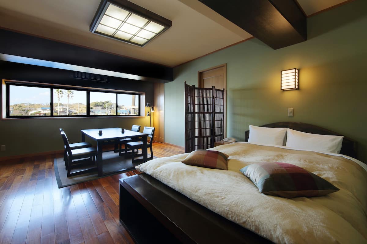 A bedroom with a large bed, a desk, and a chair. There is a window with a view of the outside.