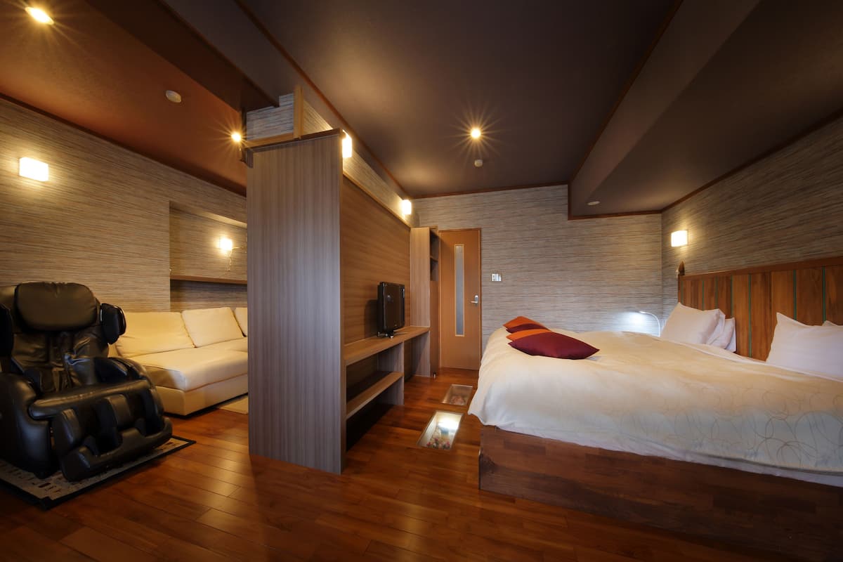 A modern hotel room with a massage chair, a sofa, a bed with white bedding, and a wooden headboard.