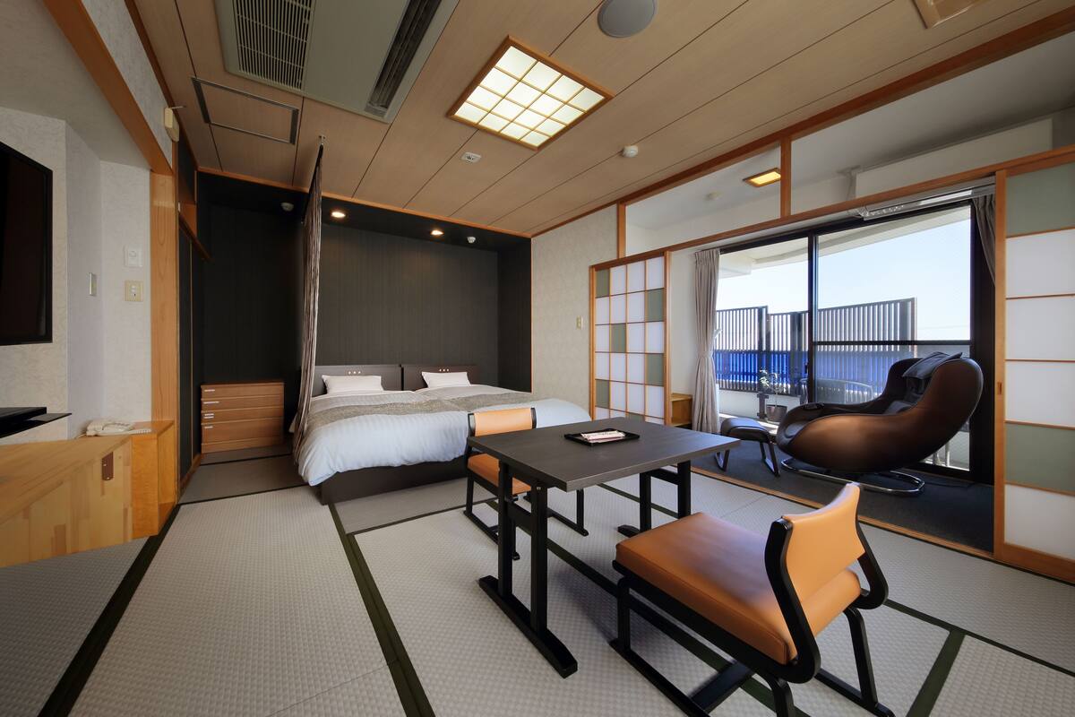 A hotel room with a bed, a desk, a chair, a sofa, a sliding door, and a view of the outside.