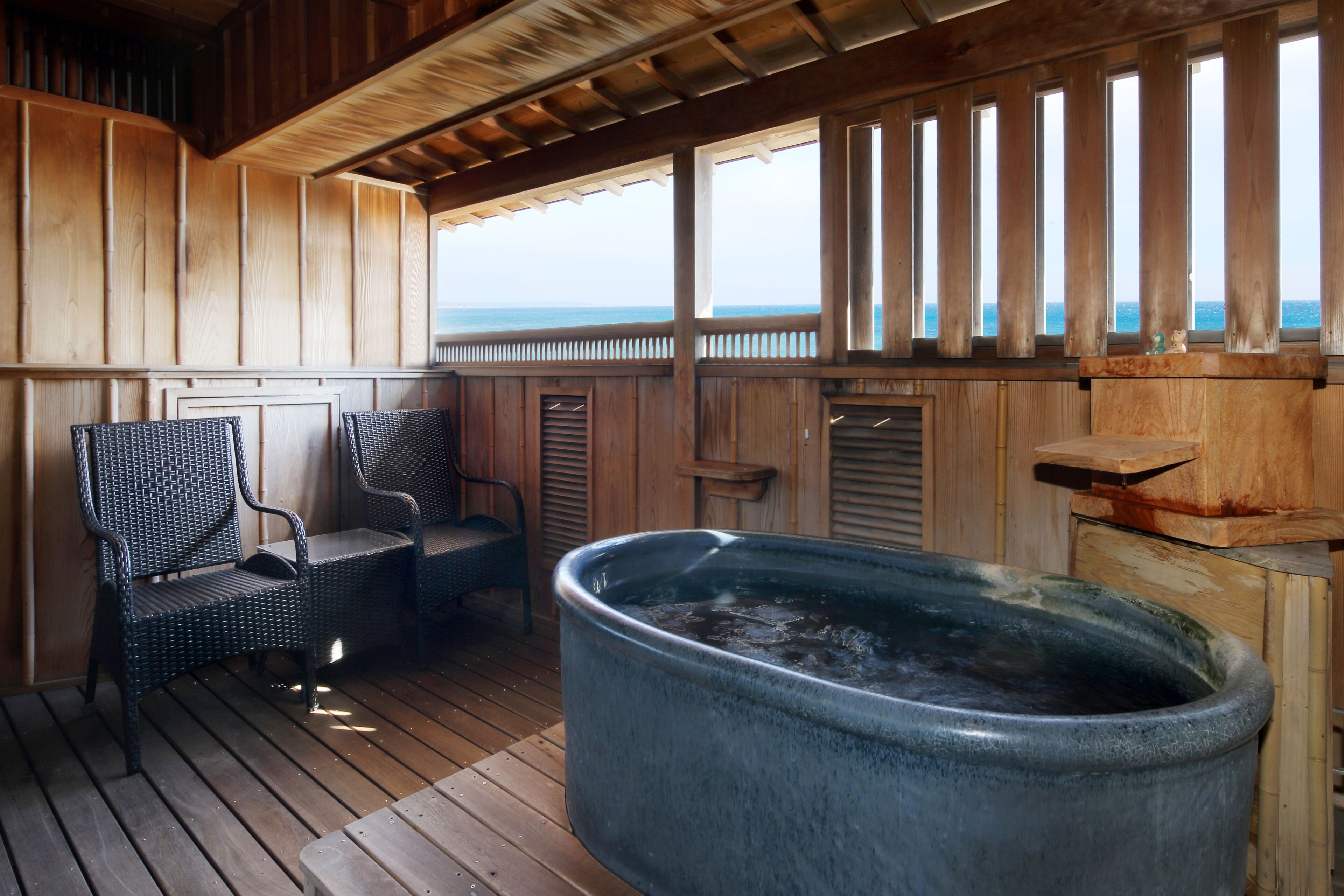 Eine Holzterrasse mit einem Whirlpool, zwei Korbstühlen und Blick auf den Ozean.