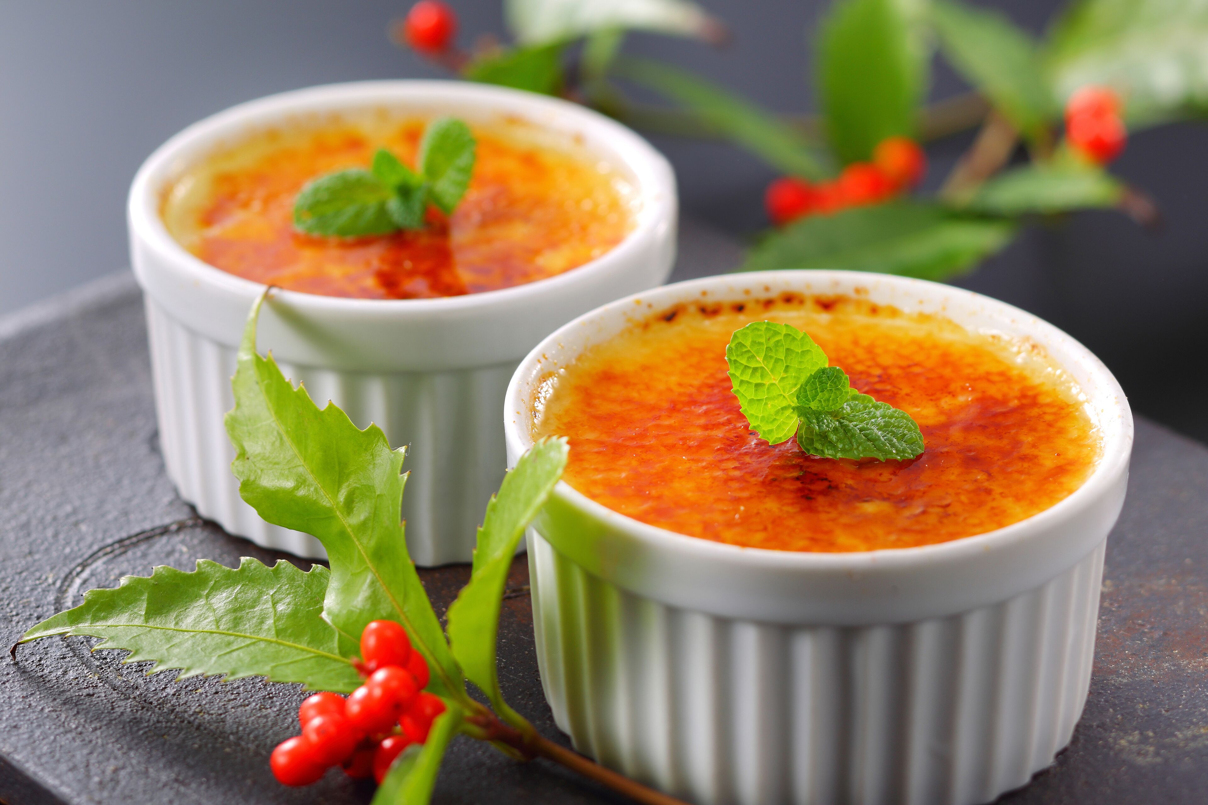 Two white ramekins of crème brûlée on a dark surface with green leaves and red berries.