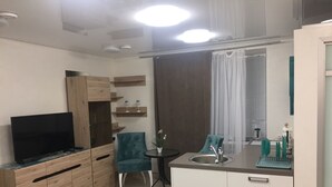 En kompakt opholdsstue med et tekøkken, et fjernsyn på et træskab, en turkis lænestol og et lille bord med en potteplante.