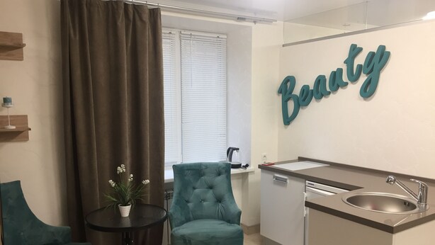 Et moderne tekøkken med vask, et lille bord og en teal farvet stol. Et stort spejl med ordet "Beauty" hænger på væggen.