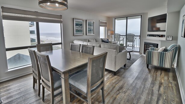 Un amplio salón con un ventanal grande con vistas a la playa, un comedor con mesa y sillas de madera, y una zona de estar acogedora con sofá y sillones.