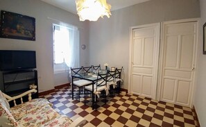 Een kamer met een zwart-wit betegelde vloer, een eettafel met stoelen, een televisie, een bank en een deur.